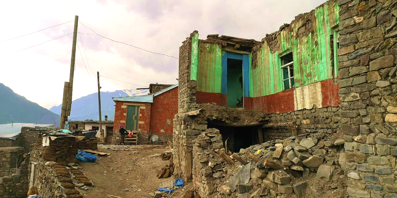 Cluster of traditional stone houses in Khinalig village on steep dirt lane, some walls brightly painted green and red while others are crumbling, exposed wooden beams and collapsed roof sections, small blue-painted doorway and window, rough stone retaining walls and scattered rubble in foreground, snow-capped mountains and cloudy sky in background