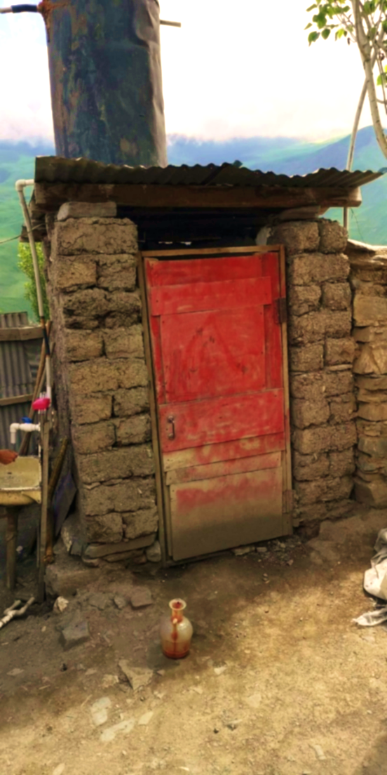 Traditional outdoor toilet structure in Khinalig village with weathered brown mud-brick walls, bright red painted wooden door, corrugated metal roof, wooden support pole, bare ground, lush green forested mountains in background, simple utilitarian construction