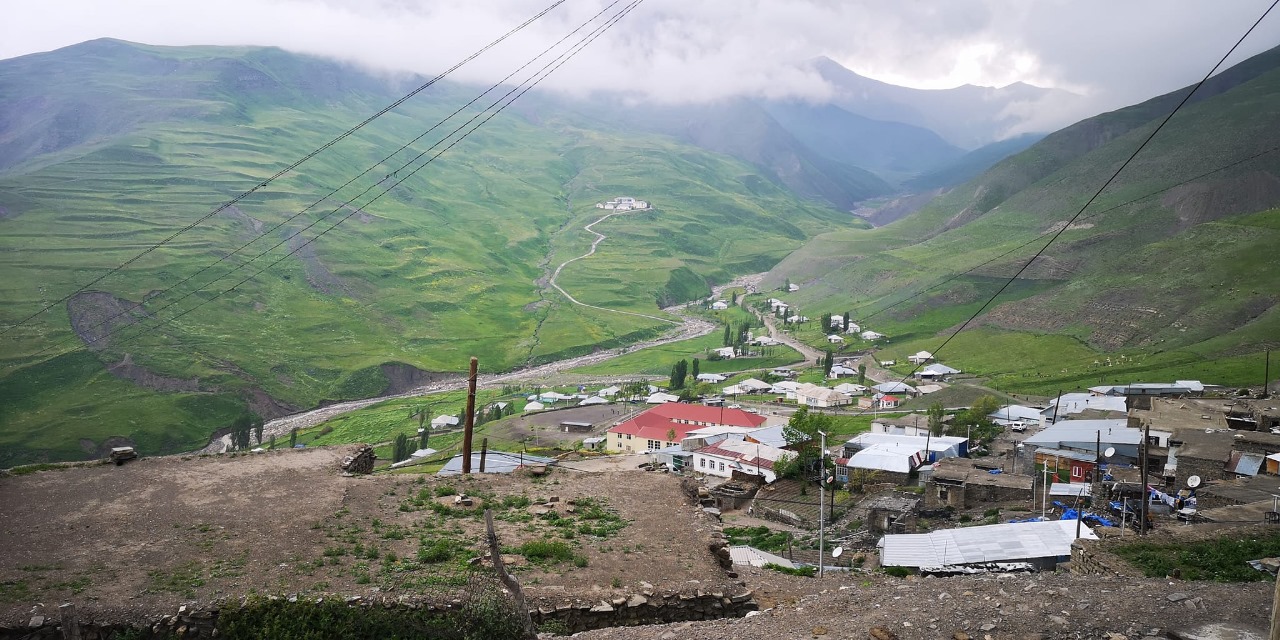 Mountain village of Khinalig in Greater Caucasus showing cluster of traditional stone houses with metal and tile roofs on terraced hillside at 2,350 meters elevation, green valley with winding dirt road cutting through rolling hills, scattered buildings along roadway, power transmission lines crossing landscape, distant mountains shrouded in low clouds and mist