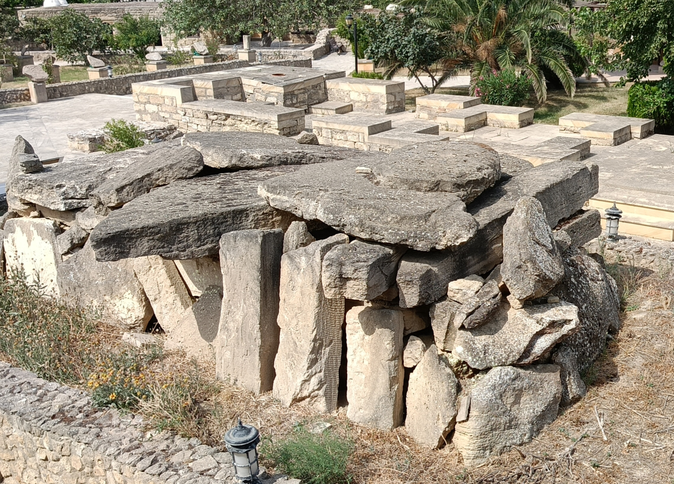 Ancient Bronze Age burial tomb structure at Gala State Historical Ethnographic Reserve showing large weathered capstones supported by vertical stone slabs creating burial chamber, with modern paved pathways and garden landscaping visible in background