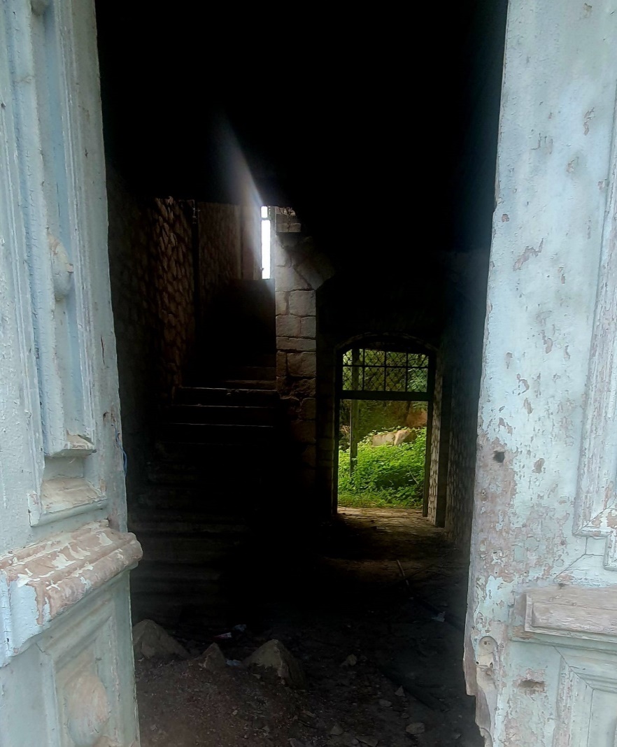 Interior view of war-damaged abandoned building in Qarabağ showing deteriorated staircase with peeling paint on pale blue walls, debris-covered floor, doorway opening to overgrown exterior greenery, and harsh natural light streaming through damaged structure