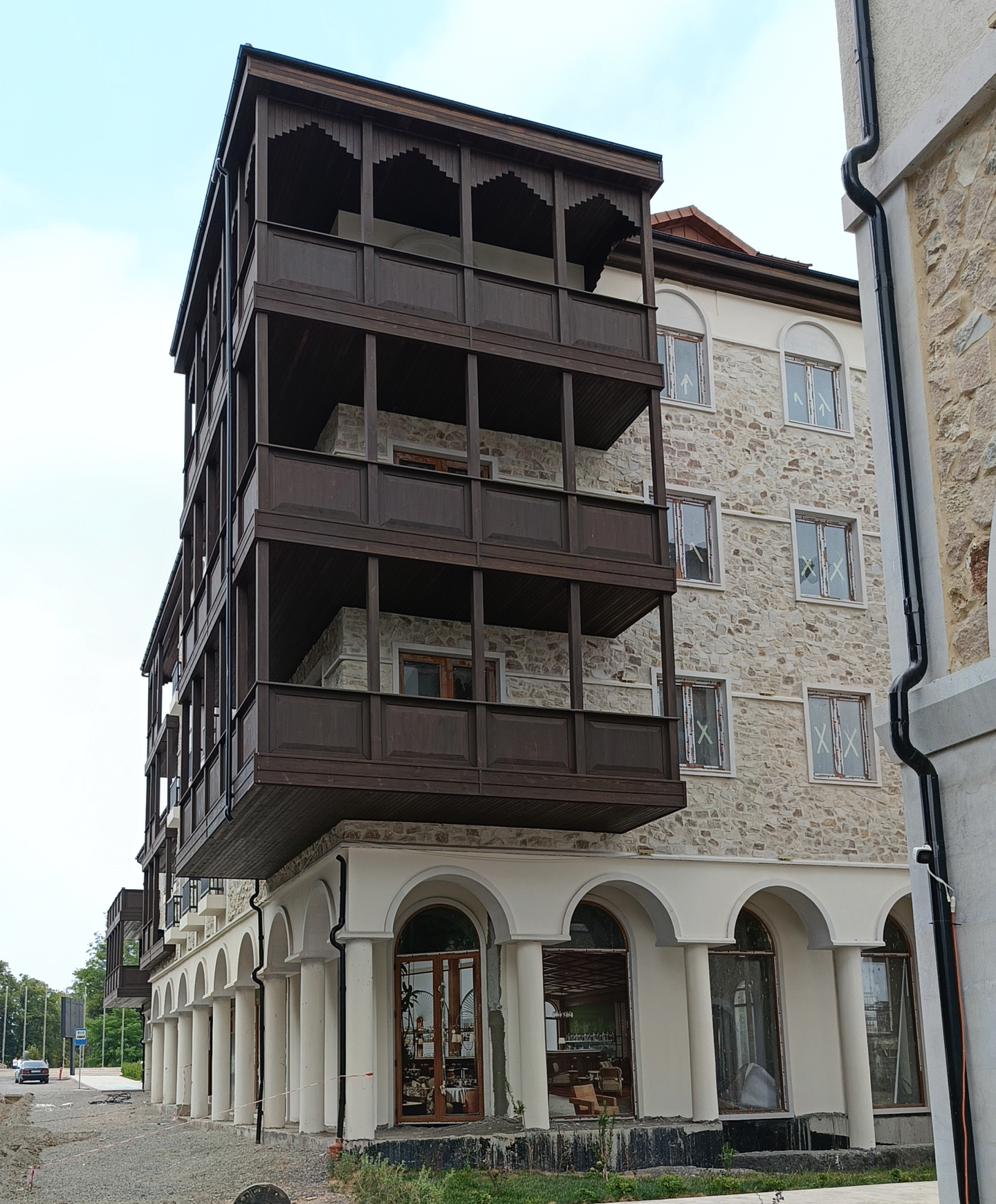 Modern multi-story building in Khankendi featuring three levels of traditional dark wooden enclosed balconies with decorative scalloped trim, projecting from stone façade with arched ground-floor arcade, white columns, and modern glass windows
