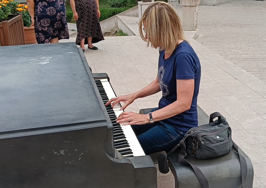 Dr. Wonda Grobbelaar seated at public piano in Khankendi park, wearing blue t-shirt and jeans, playing keyboard outdoors on paved plaza with pedestrians in background and black backpack beside bench