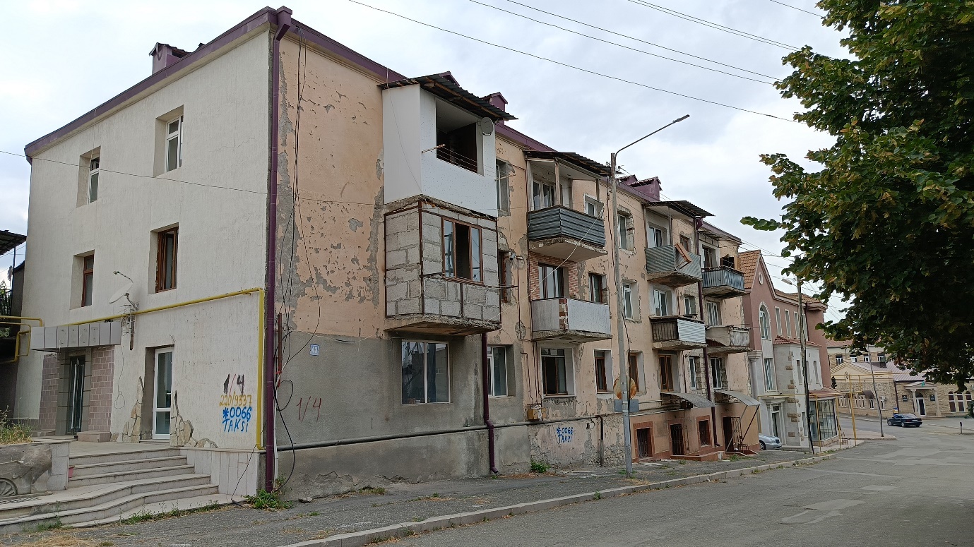 Weathered Soviet-era three-story residential apartment building in Tovuz, Azerbaijan, with peeling stucco walls in beige and pink tones, damaged roof sections, enclosed balconies with metal railings, ground-level commercial spaces with arched openings, scattered graffiti, and overgrown vegetation along quiet paved street