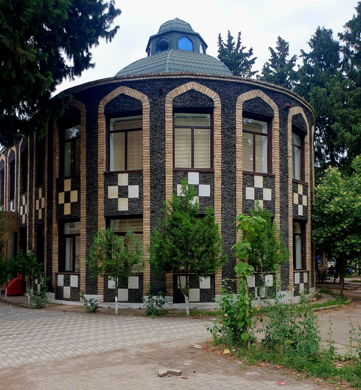 Modern circular building in Tovuz, Azerbaijan, featuring ornate brickwork in alternating dark and tan colors creating checkerboard patterns and geometric designs, with arched windows, central blue-domed cupola with windows, wooden framing above, and young trees in foreground