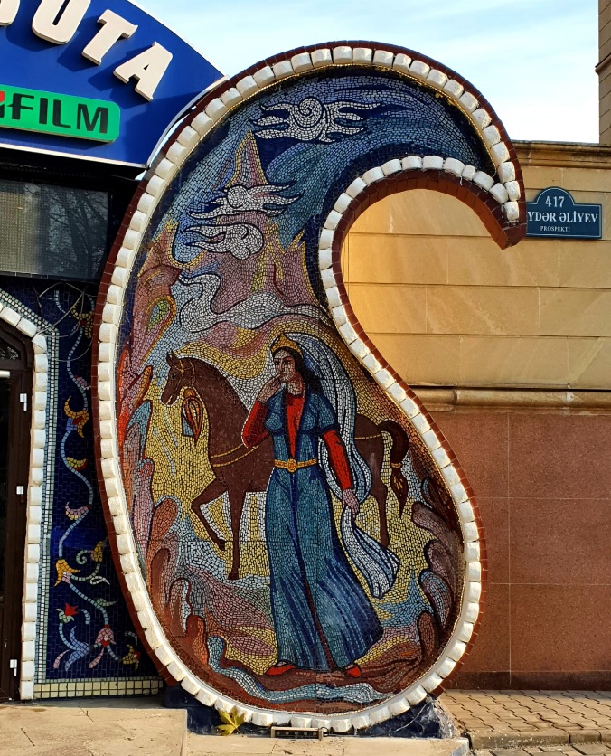 Large decorative mosaic sculpture in the shape of a traditional Azerbaijani tar (stringed musical instrument) depicting a woman in traditional dress with a brown horse, set against bare winter trees on a Ganja city street