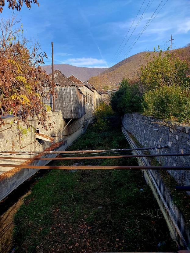 A traditional irrigation canal in Sheki, Azerbaijan, flowing between stone-walled residential properties with mountains visible in the background under a clear blue autumn sky