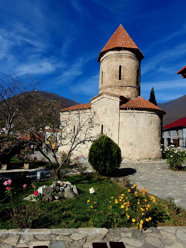 The Church of Kish in Sheki, Azerbaijan—a circular Caucasian Albanian church with distinctive red-tiled pyramidal roof, weathered limestone walls, and small window openings, set against a bright blue sky with surrounding mountain landscape and garden setting.