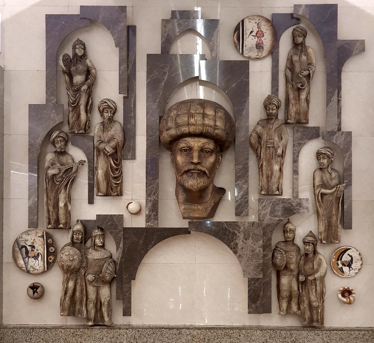 A decorative relief wall featuring Azerbaijani historical figures and cultural symbols, including a central figure wearing a traditional tall hat surrounded by various characters in traditional dress