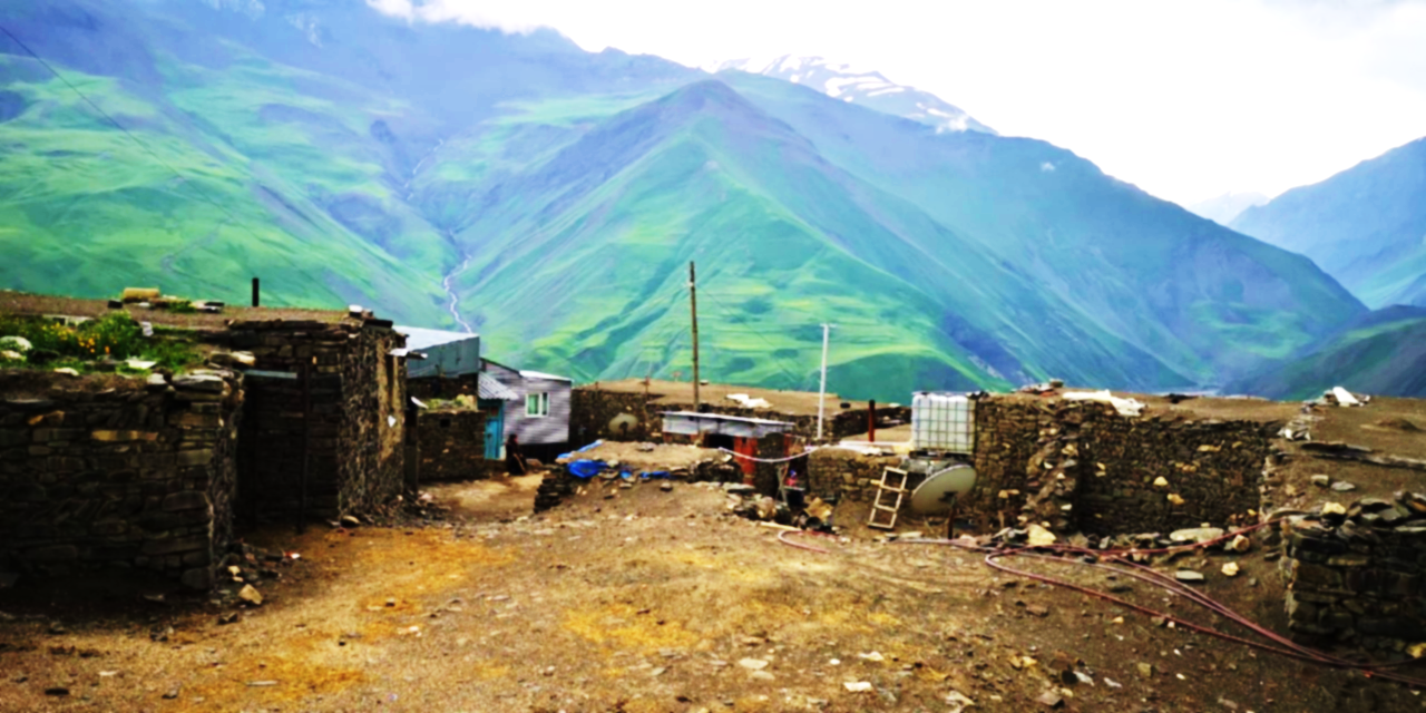 Panoramic view from Khinalig village over clustered stone houses on a high plateau, with deep green and brown valleys and a distant snow‑tipped Caucasus mountain ridge that marks the border between Azerbaijan and Russia (Dagestan)