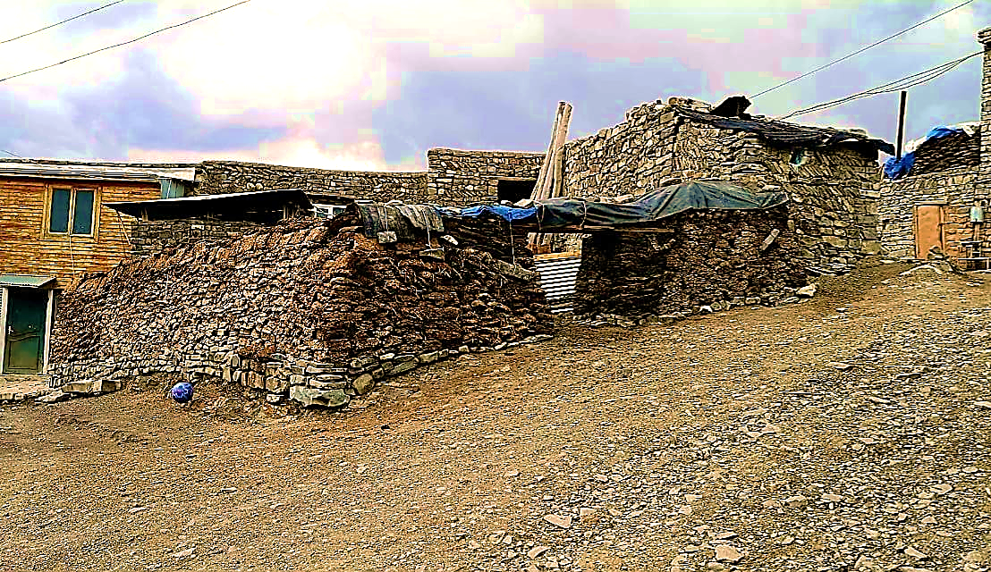 Old traditional stone house in Khinalig village with thick uneven stone walls, small wooden-framed windows, partially collapsing roof, and rough dirt foreground, perched on steep mountainside with distant Caucasus peaks in background