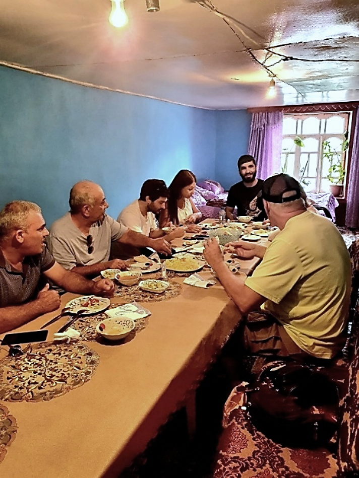 Khinalig residents and tour group (including author and husband) seated around a long dining table inside a modest home, sharing a meal with multiple small dishes,